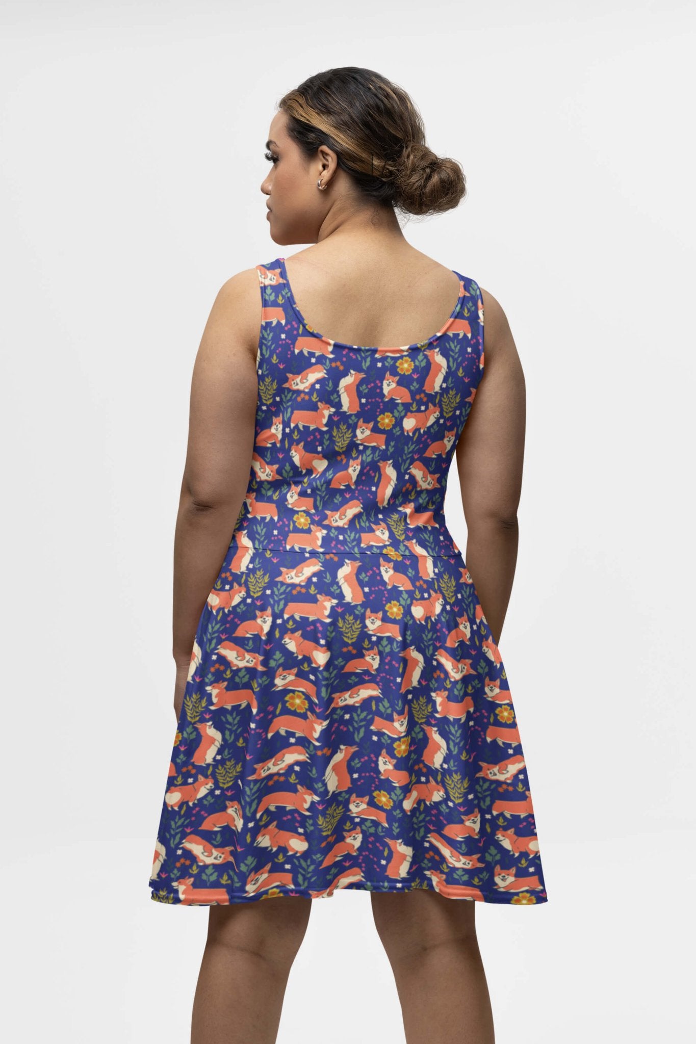 The individual sports a Cute Corgi Garden Party Skater Dress from Dogloverclothing.com, featuring a vibrant print of foxes, leaves, and other flora on a dark blue background. Their hair is styled in a low bun as they stand facing away.