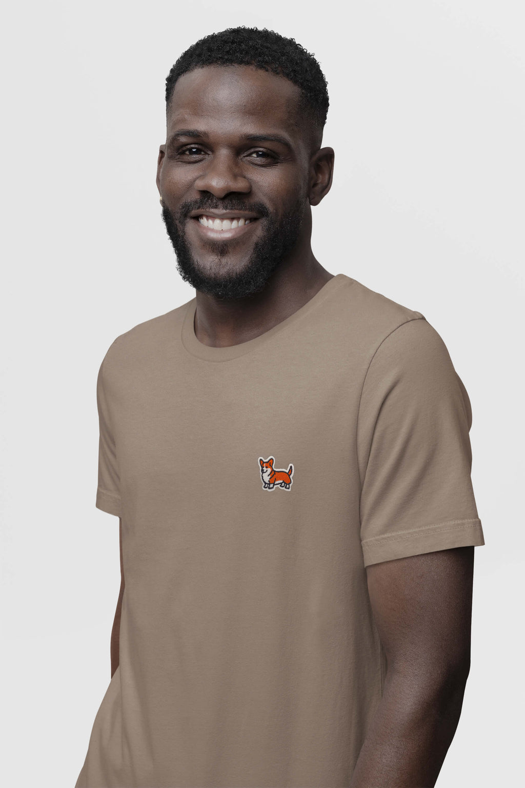 A bearded man smiles while wearing a tan Corgi Tail Wagger Unisex T-Shirt from Dogloverclothing.com, featuring a small embroidered fox on the chest against a plain white background, ideal for corgi enthusiasts.