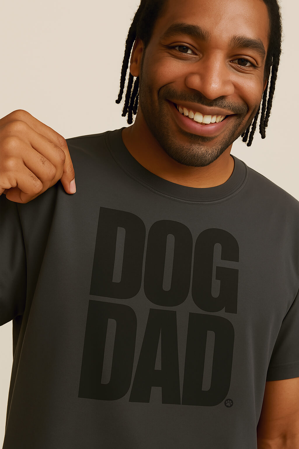 A smiling man with short dreadlocks holds his Oversized Dog Dad T-Shirt from Dogloverclothing.com. This relaxed fit, dark gray tee features bold DOG DAD lettering and is made from durable cotton for long-lasting comfort and style.