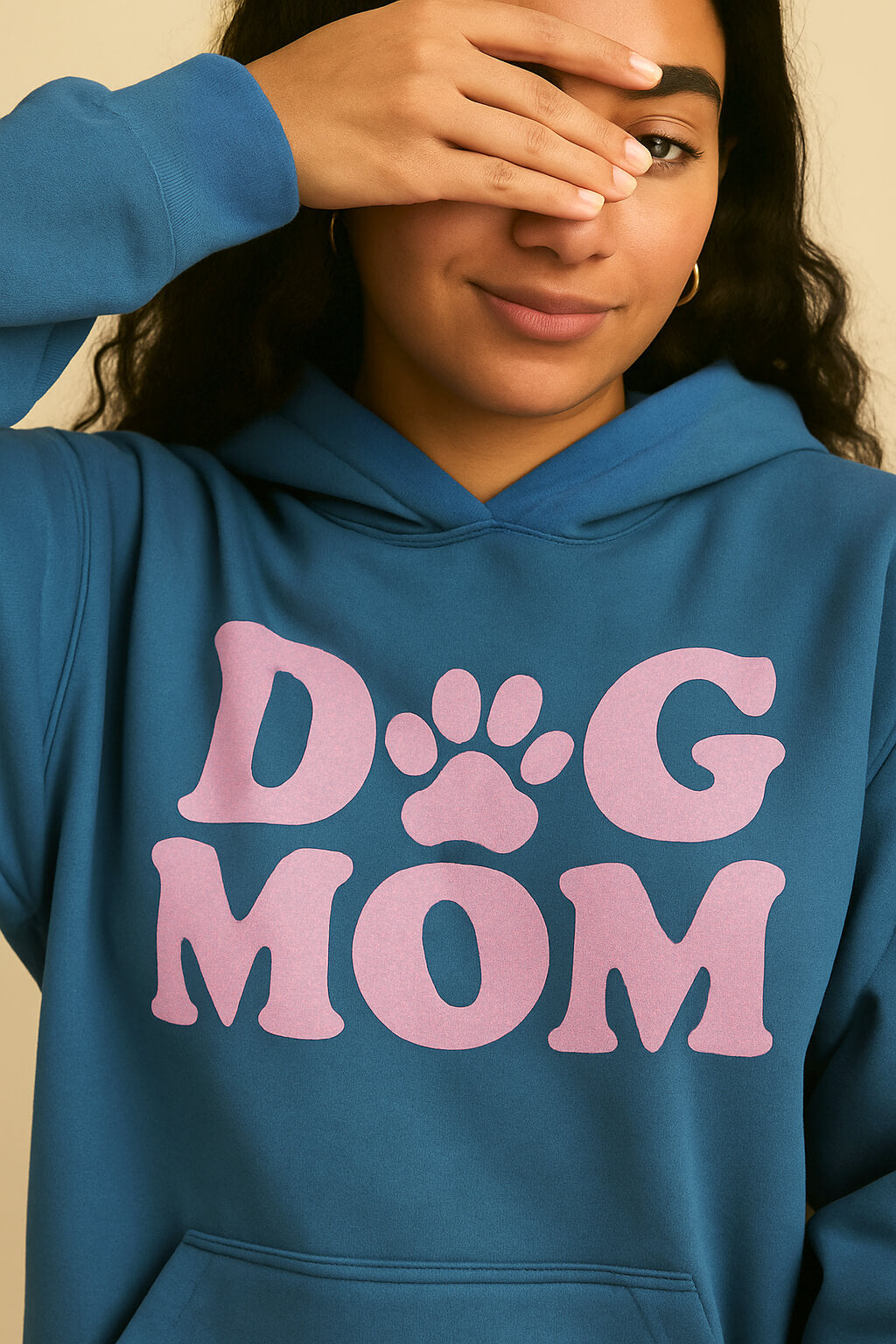 A woman wears the ultra-soft Dog Mom Hoodie from Dogloverclothing.com, featuring large pink DOG MOM letters with a paw-print O. She smiles softly, playfully covering one eye with her hand.