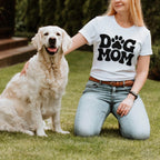 A woman in the white Dog Mom T-shirt from Dogloverclothing.com kneels on the grass beside her smiling Golden Retriever, both radiating joy amid vibrant greenery. The dogs snazzy bandana adds charm to this delightful scene for dog lover items enthusiasts.