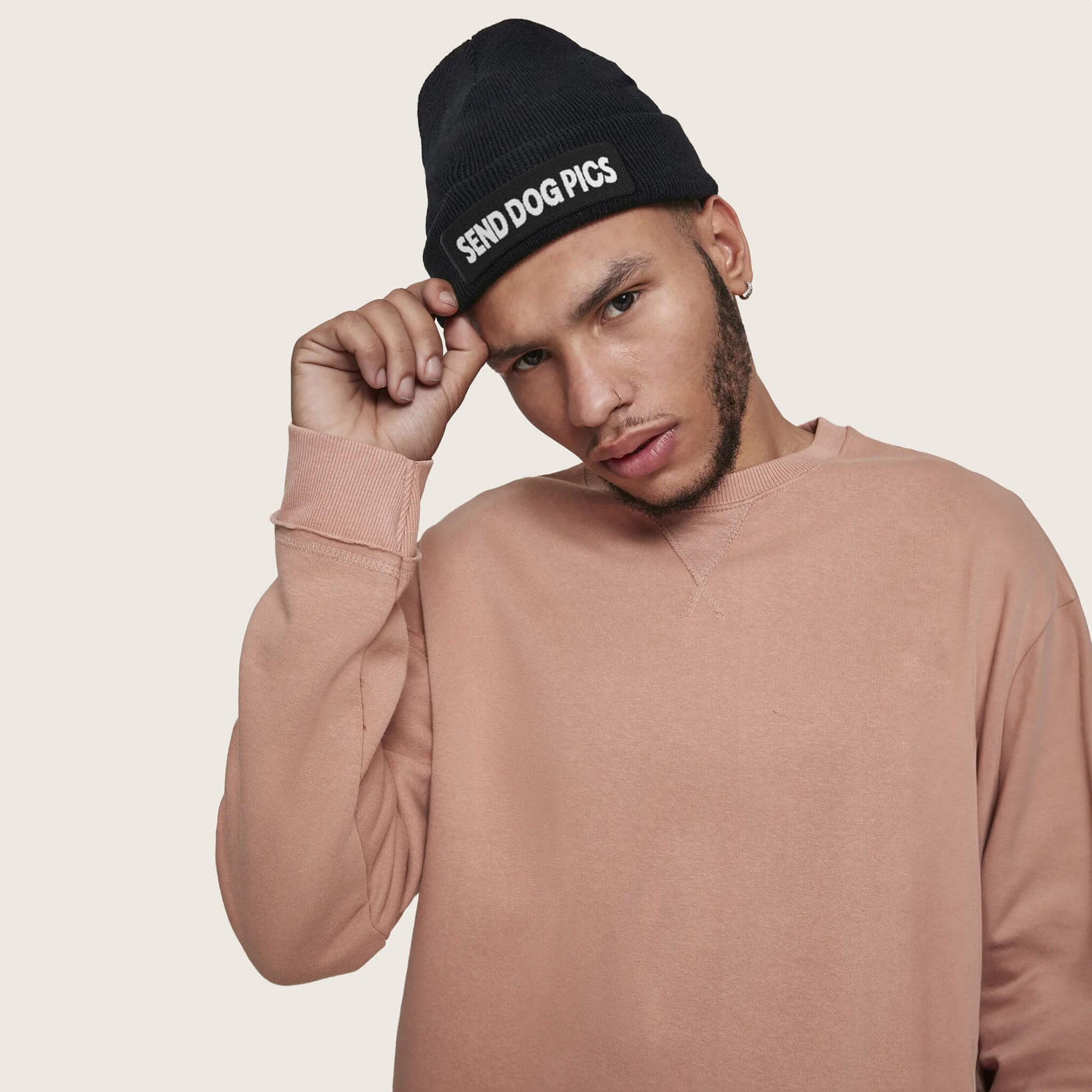 A person in a peach sweatshirt dons the Send Dog Pics Unisex Beanie from Dogloverclothing.com, standing against a plain background. With one hand on the beanie, they gaze at the camera neutrally, embodying their love for dog-themed items.