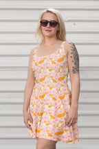 A person with long blonde hair, wearing sunglasses and the Adorable Corgi Summer Dress by Dogloverclothing.com, stands against a light-colored, corrugated metal background. They have a floral tattoo on their left arm, capturing true corgi cuteness.