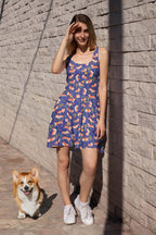 A woman wearing the Cute Corgi Garden Party Skater Dress from Dogloverclothing.com stands against a stone wall, shielding her eyes from the sun. She smiles with a corgi beside her on the pavement as shadows create diagonal lines across the scene.