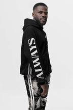 A man stands against a plain background, wearing the Corgi Supreme Tribute Hoodie—a black, eco-friendly hoodie from Dogloverclothing.com with ALWAYS on the sleeve. Paired with vertical striped pants, his attire of recycled materials complements his beard, short hair, and relaxed expression.