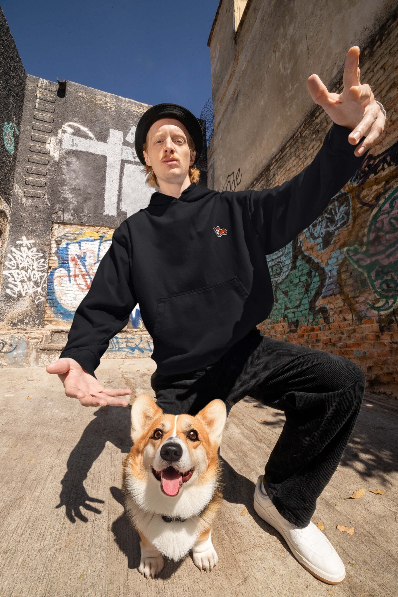 A man strikes a dramatic pose in an urban alley, wearing a Classic Corgi Sidekick Embroidered Unisex Hoodie from Dogloverclothing.com. Framed by graffiti-covered walls, a joyful corgi with its tongue out stands at his feet, highlighting the scenes maximum comfort and unabashed love.
