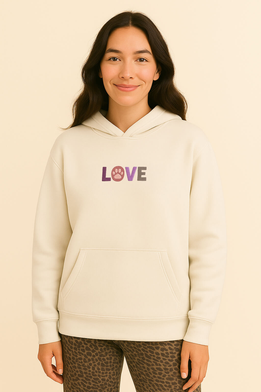 A woman with long dark hair wears the Super Soft Dog Love Hoodie by Dogloverclothing.com, featuring a cream color and embroidered LOVE design with a paw print O. She smiles against a light beige background.