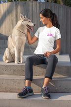 A woman in a Dogloverclothing.com Ultra Soft Dog Mama Balloon T-Shirt sits on concrete steps, smiling at her Siberian Husky. She pairs it with dark jeans and black sneakers with purple laces. The leashed dog gazes affectionately at her as sunlight casts shadows on the wall behind them.