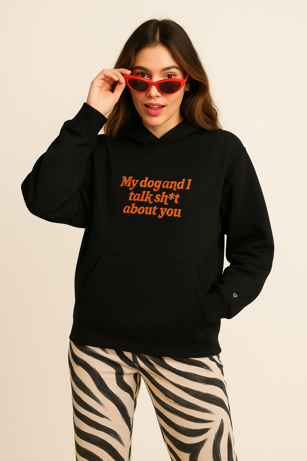 A woman in the Bold My Dog And I Talk Embroidered Hoodie by Dogloverclothing.com, with red sunglasses and zebra-print pants, stands against a beige background, lifting her sunglasses and appearing surprised.