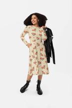 A person with curly hair wears a Cozy Corgi Long Sleeve Midi Dress by Dogloverclothing.com, paired with black boots. Holding a black jacket and smiling to the side against a plain white background, they exude comfort meets style.