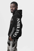 A man with a beard dons the eco-friendly Corgi Supreme Tribute Hoodie by Dogloverclothing.com, crafted from recycled materials and featuring a design with SMILING on the sleeve. He pairs it with patterned pants against a plain white backdrop.