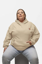 Sitting on a stool against a white backdrop, a short-haired person with hoop earrings models the Classic Corgi Sidekick Embroidered Unisex Hoodie from Dogloverclothing.com, displaying comfort and laid-back style while gazing neutrally at the camera.