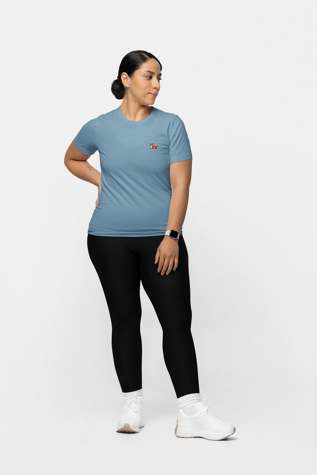 A person in a Corgi Tail Wagger Unisex T-Shirt from Dogloverclothing.com, featuring a small red logo, wears black leggings and white sneakers while standing against a plain white background. Theyre looking to the side with their hair tied back and have a watch on their wrist.