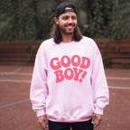 A person with long hair and a beard smiles outdoors on a tennis court, donning a backward black cap and a pink Good Boy Sweater from Dogloverclothing.com featuring bold red GOOD BOY! letters, against a blurred green background.