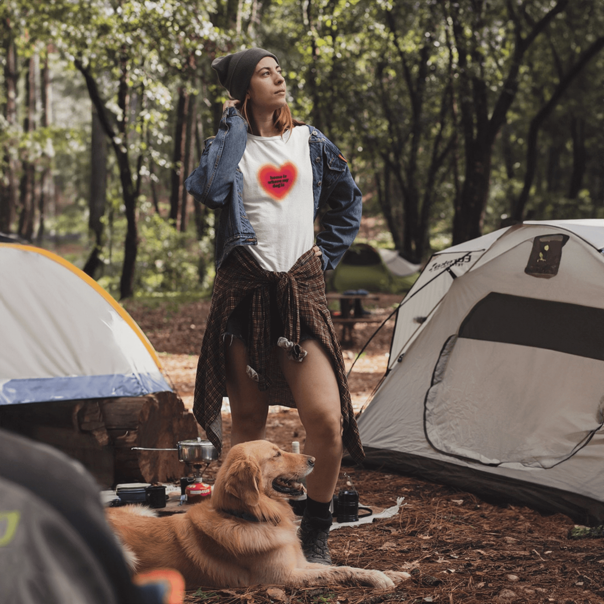 A woman stands confidently at a campsite in a denim jacket, plaid shirt tied at her waist, and the Home is Where My Dog Is unisex T-shirt from Dogloverclothing.com. A golden retriever rests on the ground nearby.
