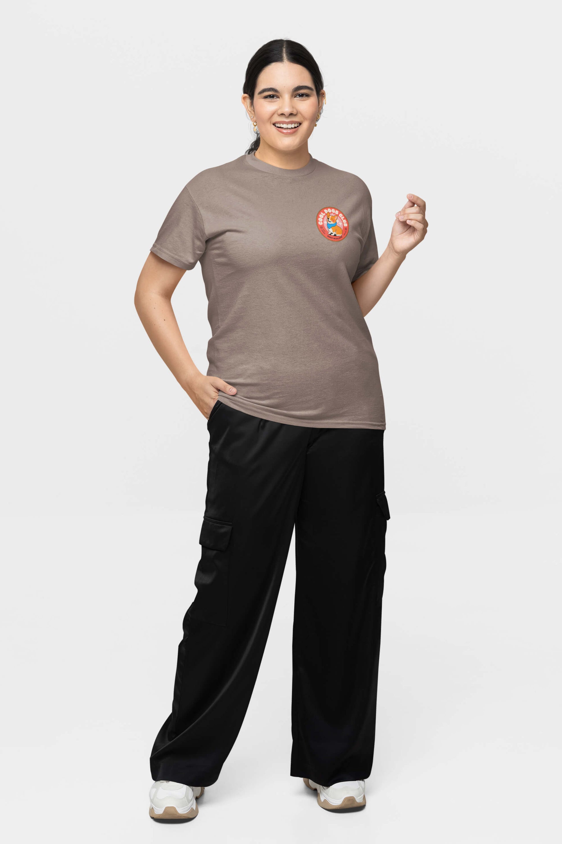 A person stands smiling against a white background, wearing the Cool Dogs Club Unisex Hype T-Shirt from Dogloverclothing.com featuring a colorful logo on the left chest, black cargo pants, and white sneakers. With their left hand in a pocket and holding an object in their right, its perfect for dog lovers.