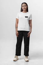 Facing forward against a plain background, the person wears a white Corgi Mom Ultimate Tribute T-Shirt from Dogloverclothing.com featuring black text on the chest and made of premium fabric. They pair it with black pants and white sneakers. Their long hair cascades past tattooed forearms, perfect for dog lovers.