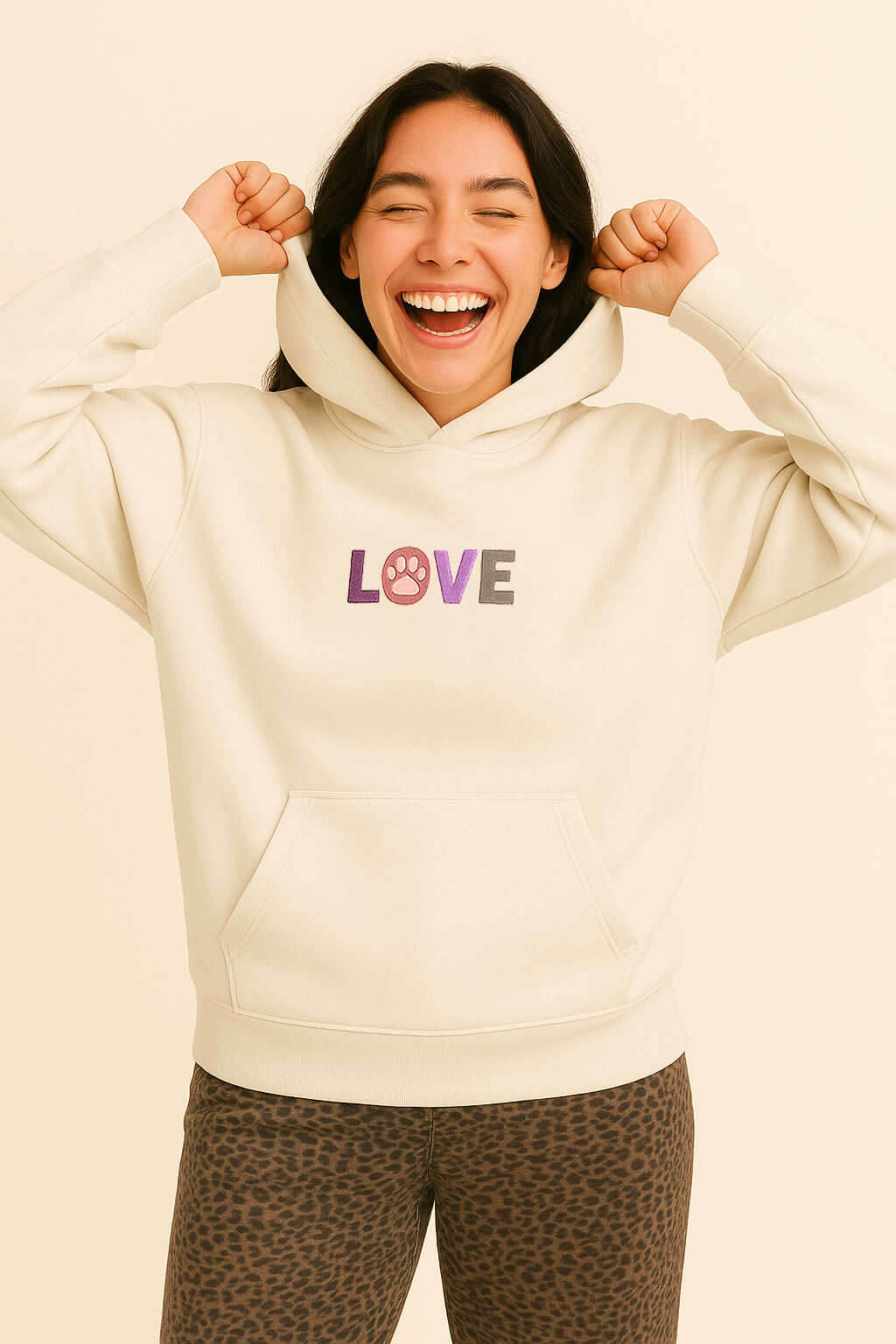 A smiling person wears the Super Soft Dog Love Hoodie from Dogloverclothing.com, featuring a cream color and embroidered LOVE with a paw print O, paired with animal print pants and showcasing fun dog parent style.