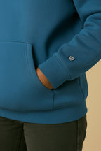 A person wears the Dog Mom Hoodie from Dogloverclothing.com in ultra-soft teal blue, featuring a small embroidered heart near the wrist and a front pocket, paired with dark pants.
