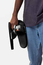 A person in blue jeans and a dark shirt holds the Mighty Corgi Rider Slides from Dogloverclothing.com, black slides with LOW and a cartoon dog on the straps—ideal for fans of comfortable footwear.