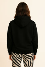 A person with long brown hair is facing away from the camera, wearing the Bold My Dog And I Talk Embroidered Hoodie from Dogloverclothing.com and black-and-white zebra print pants, against a plain light background.