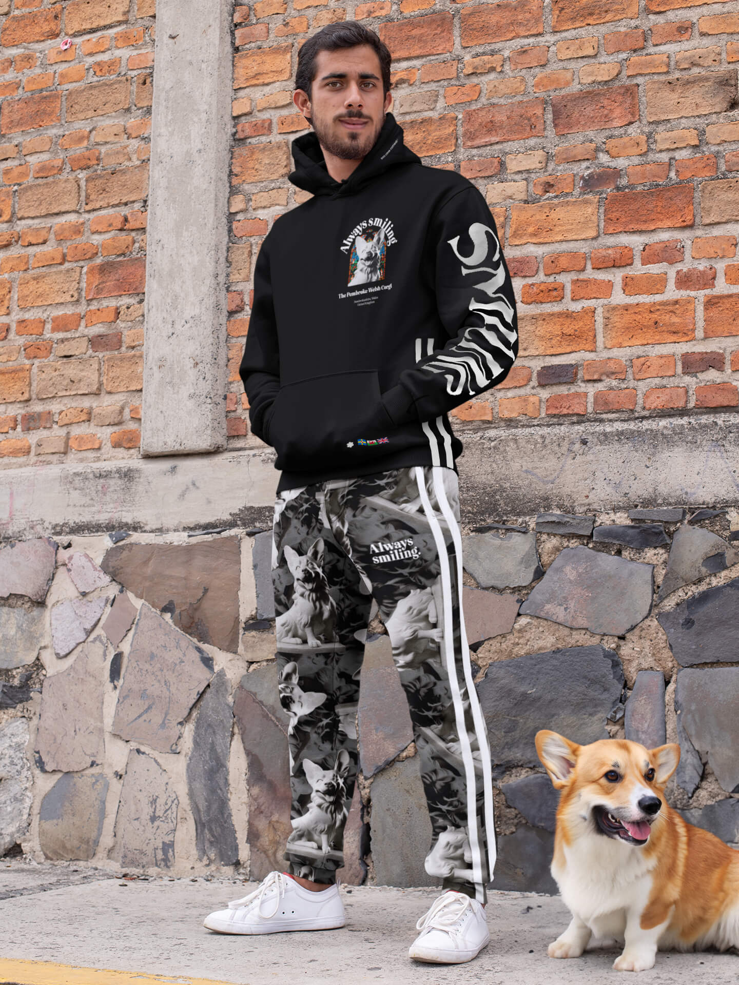 A man stands against a brick wall wearing Dogloverclothing.coms sleek black Corgi Supreme Tribute Hoodie made from recycled materials, paired with camouflage pants featuring white stripes. His hands are in his pockets as he is joined by a smiling Corgi on the pavement.