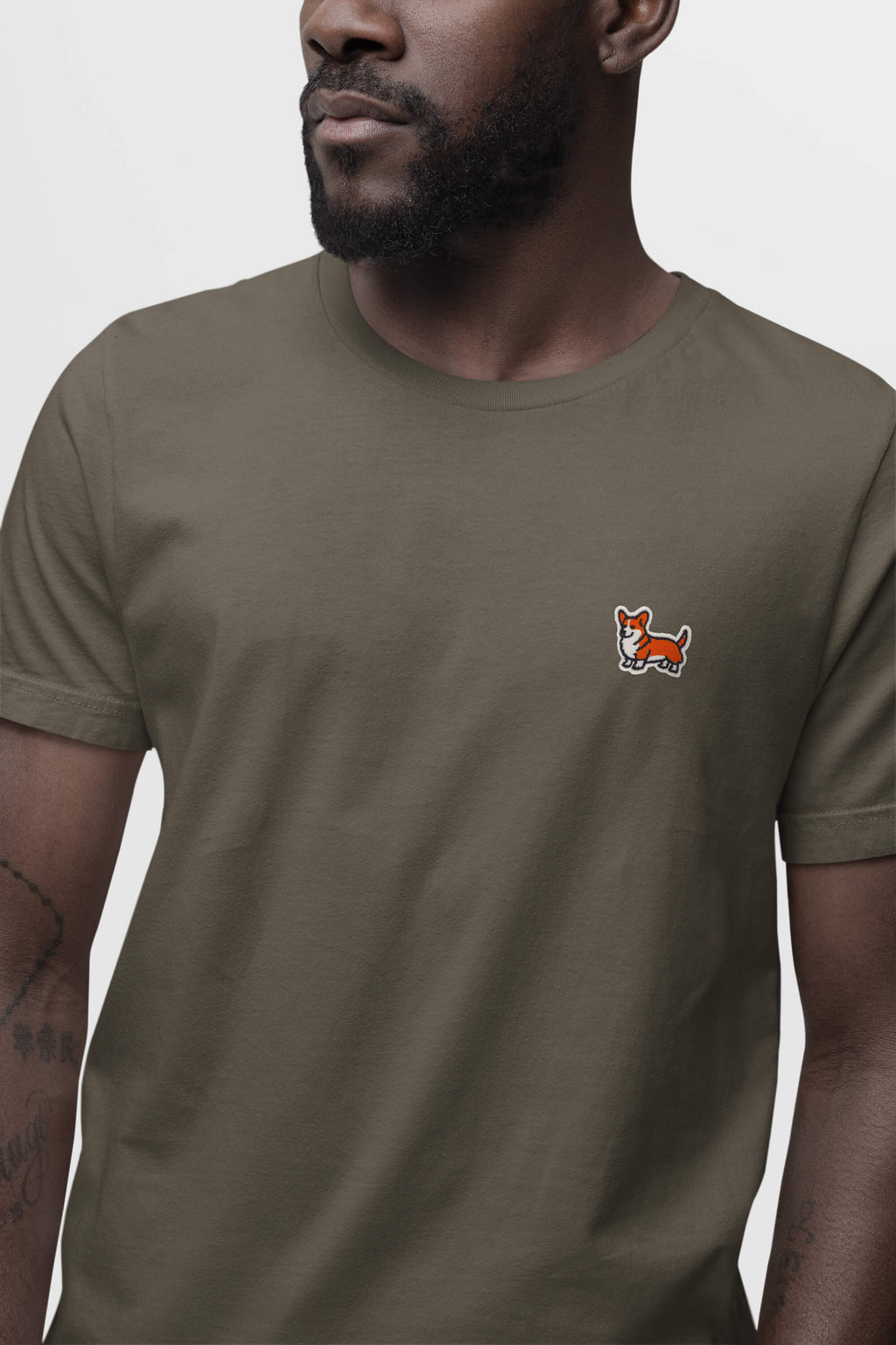 A man wears an olive green Corgi Tail Wagger Unisex T-Shirt from Dogloverclothing.com, featuring a small embroidered fox logo on the left chest. The short sleeves highlight his tattooed forearm against a plain white background, perfect for Corgi fans.