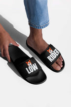 A person in blue jeans slips on Dogloverclothing.coms Mighty Corgi Rider Slides—black sandals made from durable materials, each featuring a cartoon corgi face and bold white LOW and RIDER lettering.