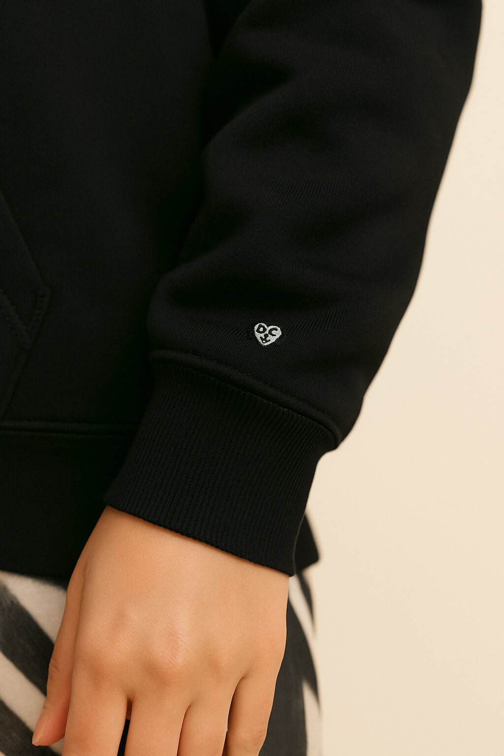 A close-up of a persons hand and lower arm wearing the Bold My Dog And I Talk Embroidered Hoodie by Dogloverclothing.com, featuring a small heart on the cuff; neutral background with classic fit black-and-white patterned pants partially visible.