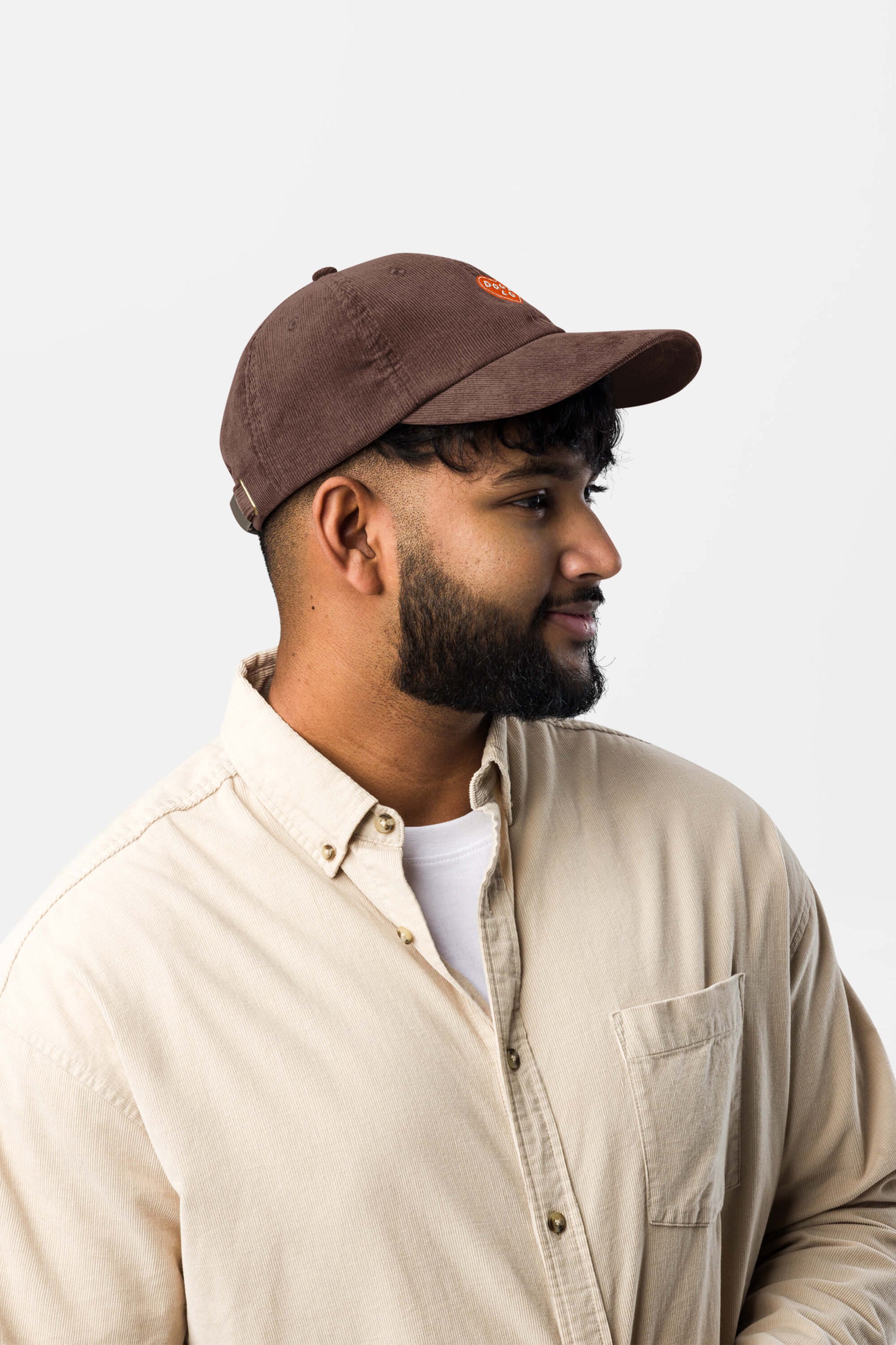 A young man with a trimmed beard wears the Limited Dog Lover Corduroy Cap by Dogloverclothing.com and a beige button-up over a white t-shirt, looking to the right against a plain light background.