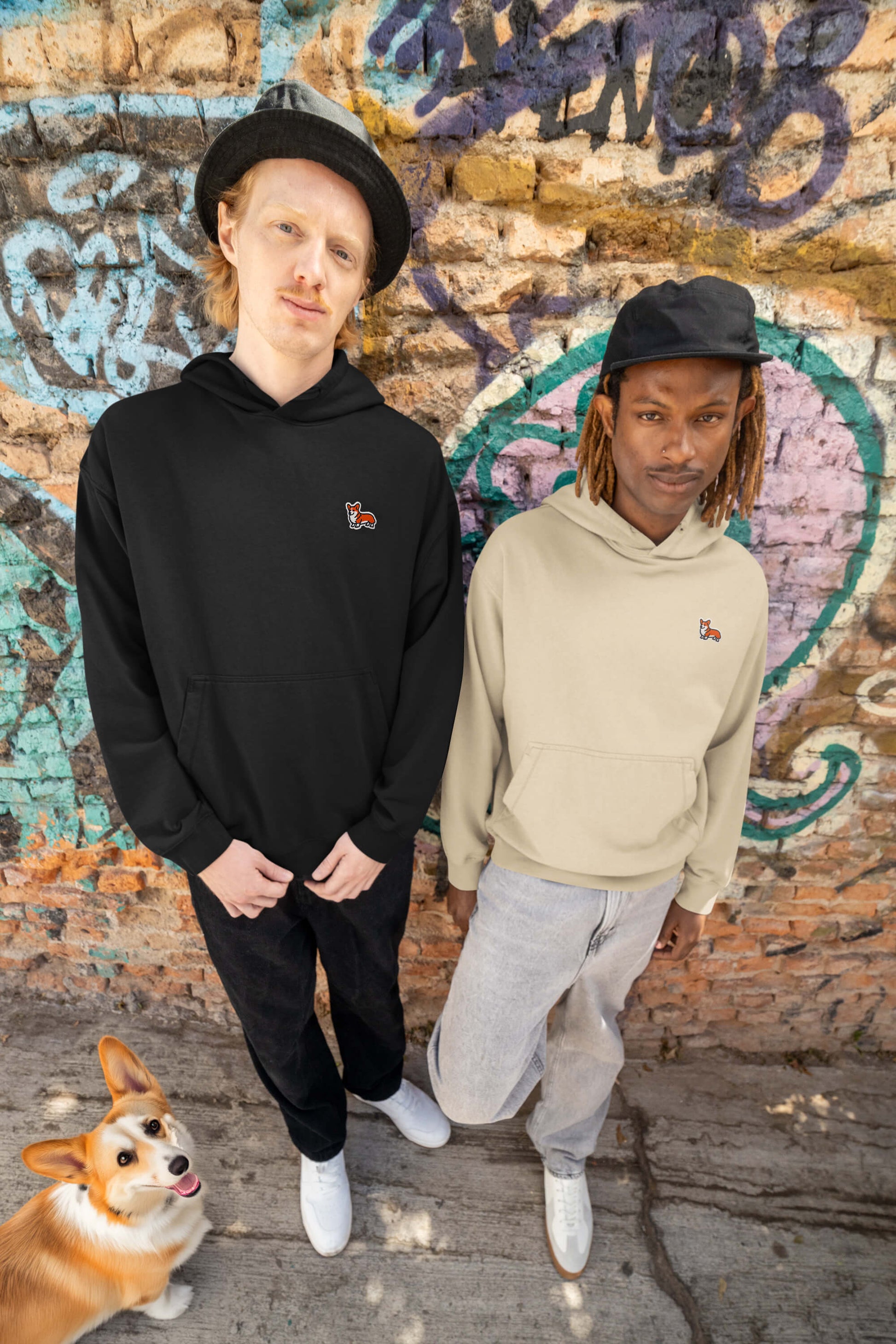 Two individuals in casual hoodies—one Classic Corgi Sidekick Embroidered Unisex from Dogloverclothing.com—stand beside a graffiti wall, wearing hats and white sneakers. A smiling corgi at their feet looks up lovingly, capturing the essence of pure joy.