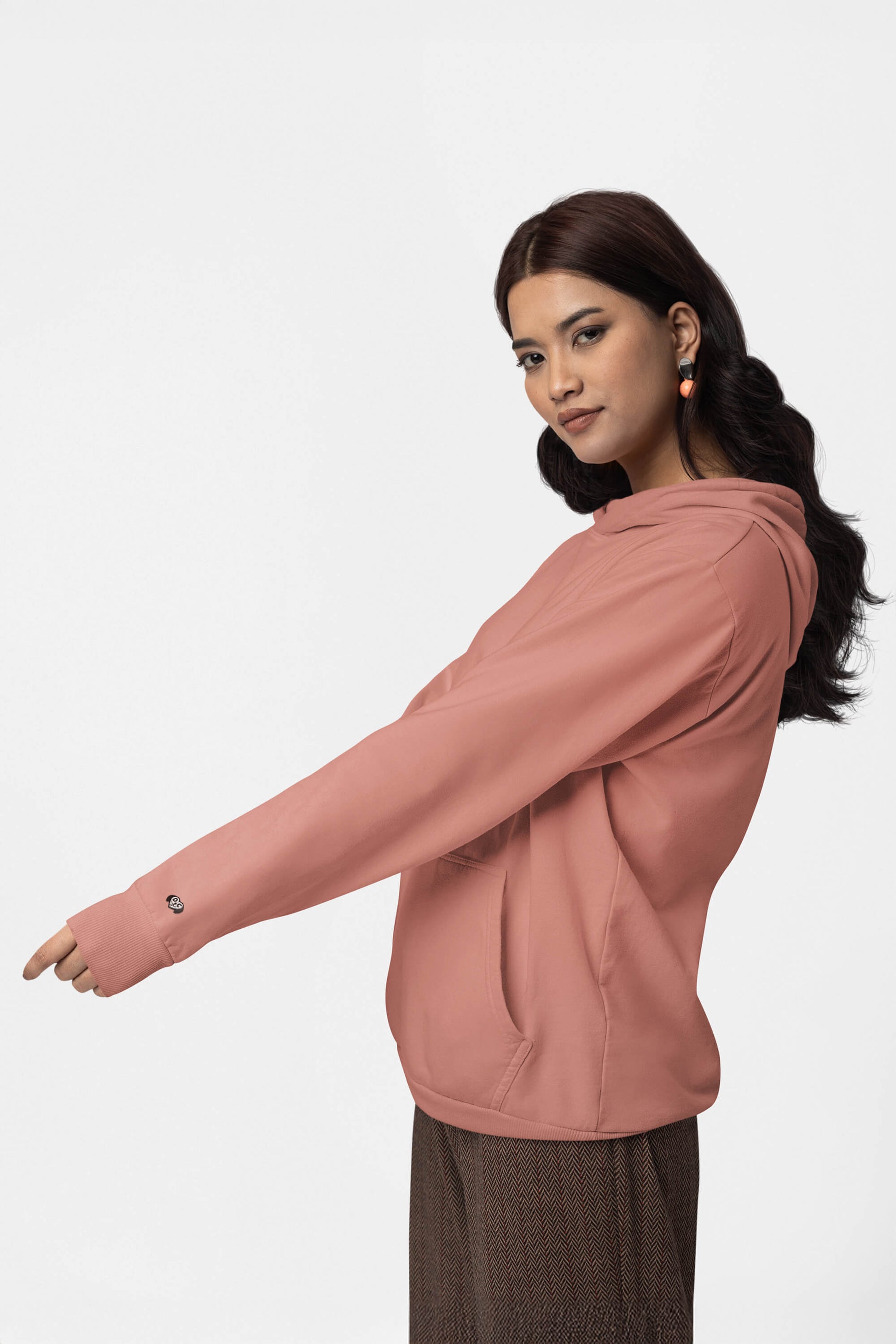 A person with long hair wears a Unisex Dog Walker© Premium Hoodie by Dogloverclothing.com in salmon, paired with checkered pants. They stand in profile against a plain white background, embodying everyday adventures and looking at the camera with a subtle smile.