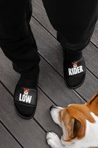 A person wearing comfortable black Mighty Corgi Rider Slides from Dogloverclothing.com stands on a wooden floor beside a brown and white dog lying down.
