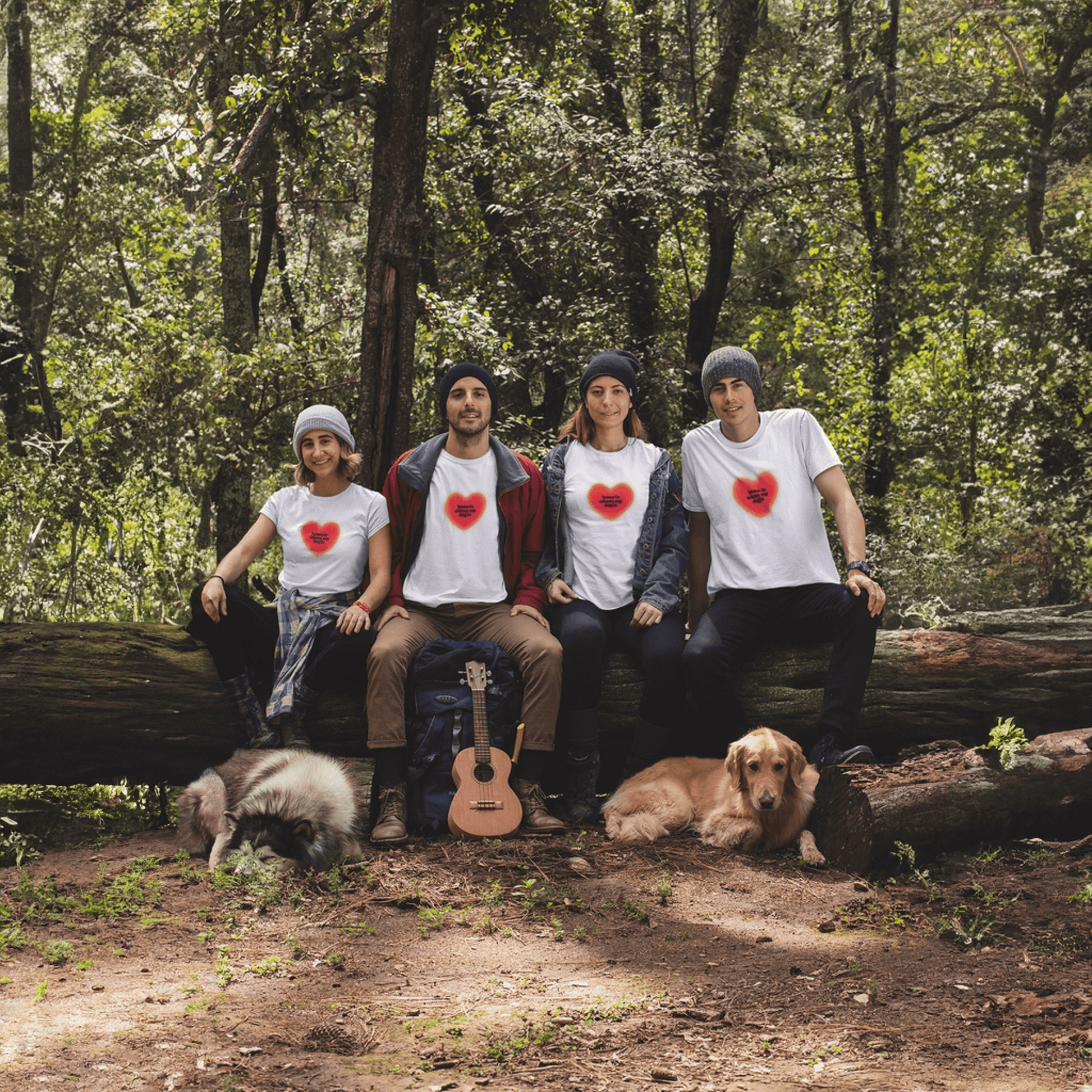Four people in Home is Where My Dog Is unisex t-shirts from Dogloverclothing.com, featuring red heart designs, sit on a forest log with two dogs. A ukulele rests nearby as they smile and enjoy the outdoors in matching premium shirts.