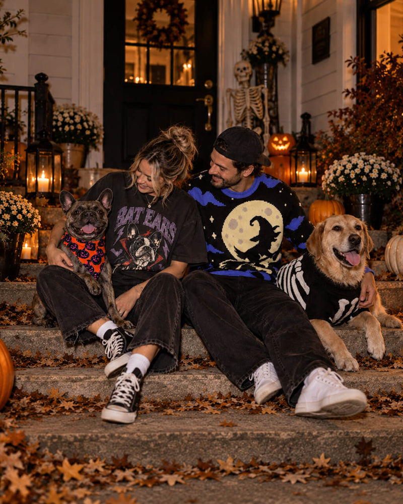 Dog parents on a Halloween porch in a Bat Ears Frenchie tee and witch sweater, with a Frenchie in a pumpkin bandana and a Golden in a skeleton sweater