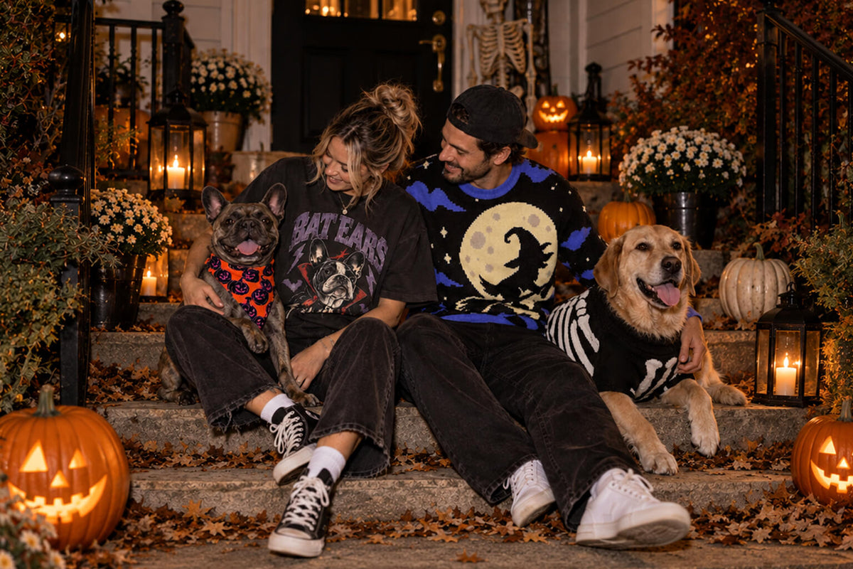Dog parents on a Halloween porch in a Bat Ears Frenchie tee and witch sweater, with a Frenchie in a pumpkin bandana and a Golden in a skeleton sweater