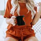 A person with long blonde hair sits on a bed wearing a short-sleeved rust outfit, holding a Dogloverclothing.com Dog Lover Mug. The cozy interior, featuring a patterned pillow and dog lover decor, enhances the rooms charm.