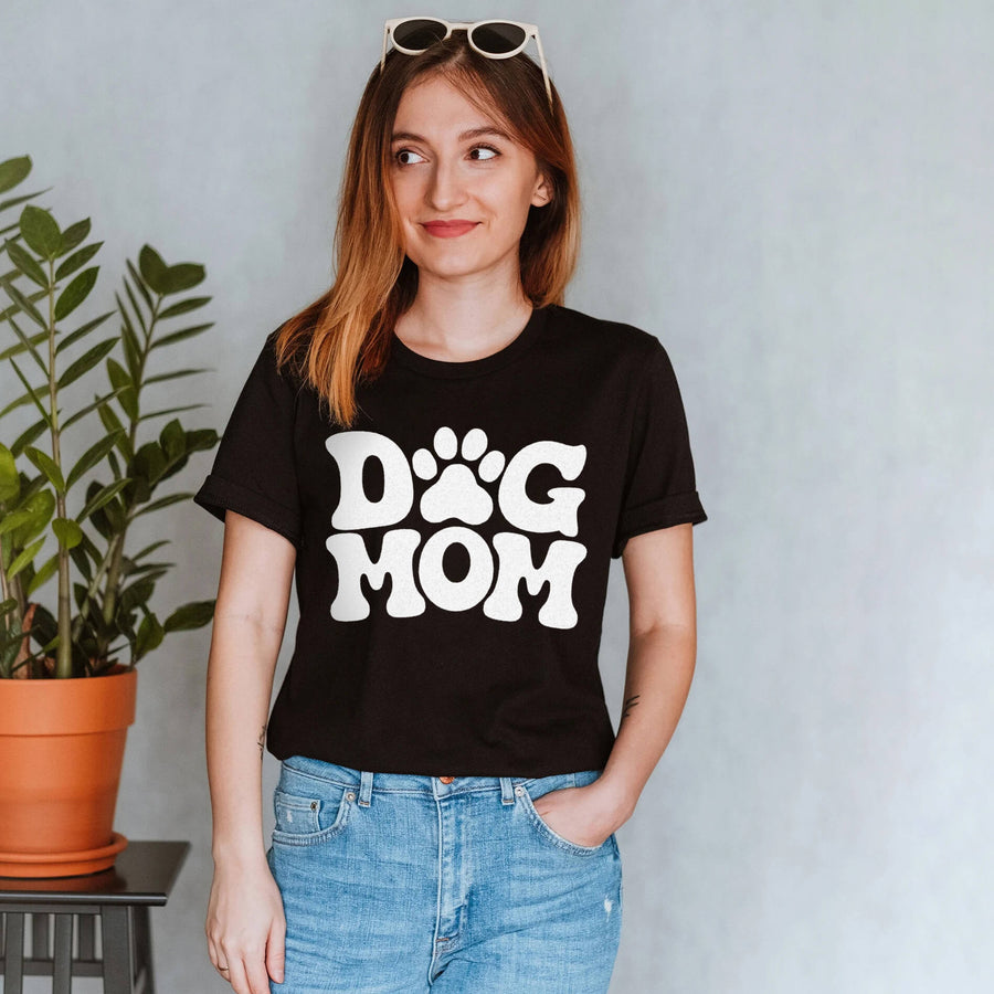 Smiling woman in a Dog Mom T-Shirt from WOOF with sunglasses on her head, standing beside a potted plant