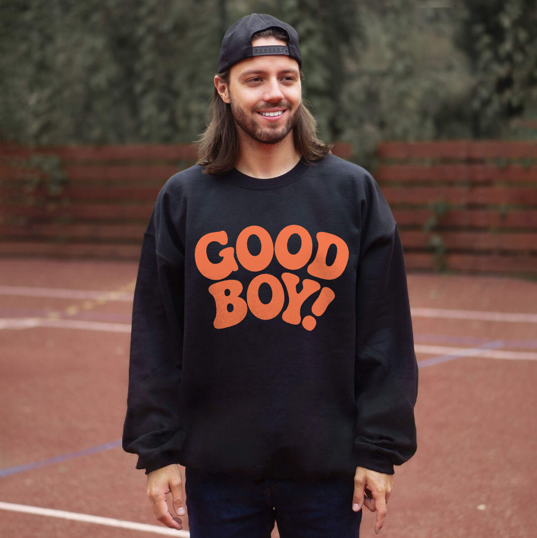 A man with long hair and a cap, embodying a true dog lover, smiles on a sports court. He wears the Good Boy Sweater from Dogloverclothing.com, featuring bold orange letters on black. A wooden fence and lush greenery enhance this charming scene.