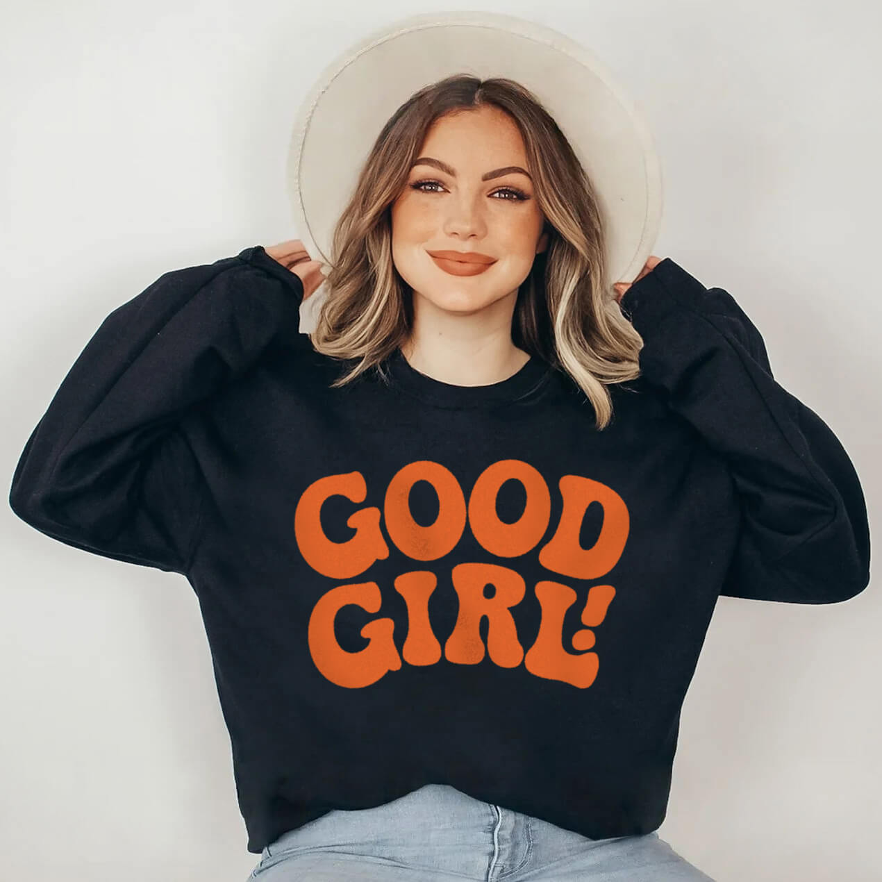 A woman with shoulder-length hair smiles, wearing a wide-brimmed hat and a black Good Girl Sweatshirt from Dogloverclothing.com that has bold orange letters. A true dog lover, she sits with her arms slightly raised against a plain background.